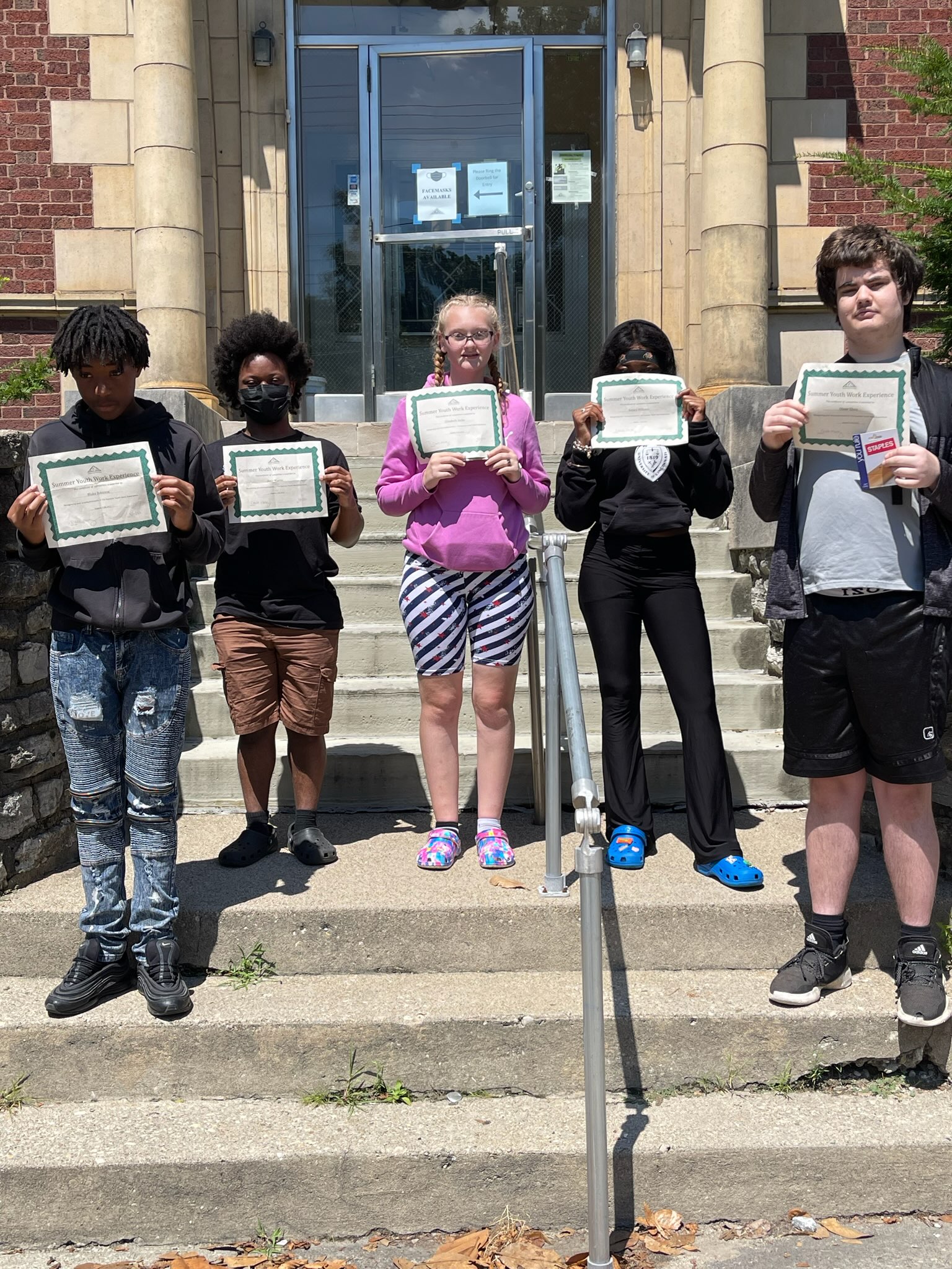 Five teenagers, each holding up a certificate of completion, stand on the steps at the IKRON building.